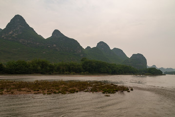 Guilin, China - May 10, 2010: Along Li River. Landscape of karst mountain under rainy cloudscape with green belt of trees on shoreline and dirt island up front.