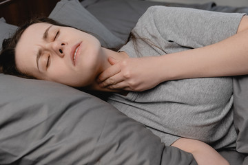 Sick upset girl lying down on bed suffering from throat sore, touching neck, having discomfort or painful feeling, sad female suffering from angina or tonsillitis. Painful swallowing, illness concept