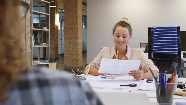 Young cheerful caucasian woman in casual clothes smiling and discussing interior design sketch with female colleague while working at desk in modern open space office