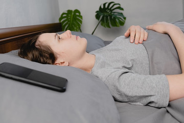 Young frustrated woman lying down on bed with phone looks into the ceiling, waiting message feels...