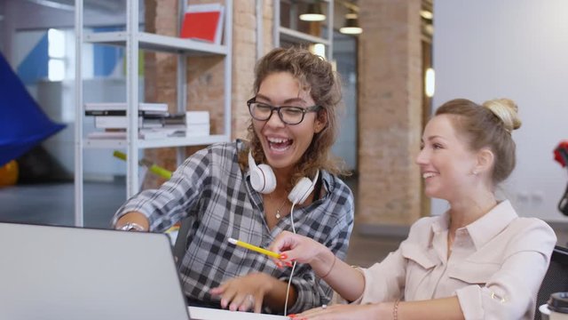 Young beautiful Caucasian and mixed raced female colleagues sitting at desk, pointing at laptop screen and laughing during workday in modern loft office