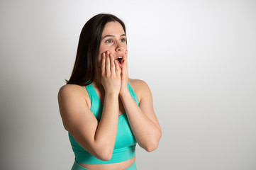 Surprised charming young brunette woman in turquoise sports clothing. Emotional expression of female athlete model isolated over white background. Active and healthy lifestyle, emotions concept...
