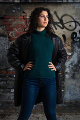 Self-confident attractive brunette in jeans and leather coat posing with her hands on hips. Female model standing at old dirty wall with graffiti...