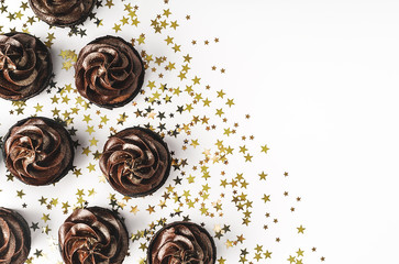 Chocolate cupcakes with chocolate icing and sprinkled gold sparkles on white background with golden star shaped confetti. Holiday concept. Mockup for your design. Flat lay style. Place for text.