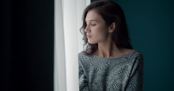 Pretty pensive girl with brown hair standing near window and looking at camera with dramatic mood. Young girl in casual clothing lost in her thoughts at home.