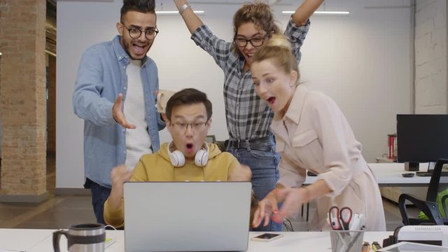 Young multiethnic colleagues reading good news on laptop screen, yelling in excitement, embracing and giving high five while celebrating business achievement in office