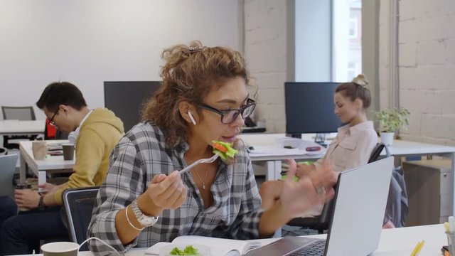 Young beautiful mixed raced woman in wireless headphones and casualwear sitting at desk, eating salad, talking via online call on laptop and taking notes while working in modern coworking space