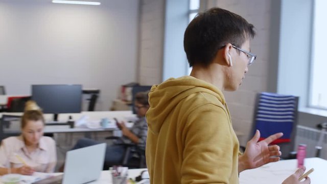 Young Asian man in wireless headphones and casual clothes walking in coworking space and talking via speakerphone while colleagues working in background