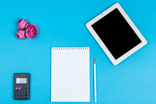 top view of the workspace on a blue background. calculator, a notebook, a pencil, a tablet computer and pink flower buds