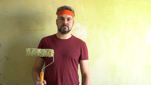 A bearded man is standing against the wall. In the hand is the roller. On the head is an orange ribbon. Repair work in the room. Frick. Retro style. Green wall.