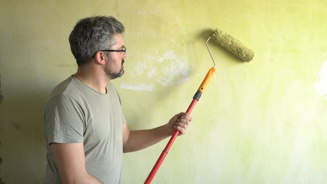 A bearded man paints a wall. Good mood. A smile on his face. Repair work in the room. Actor. Frick. Clown.
