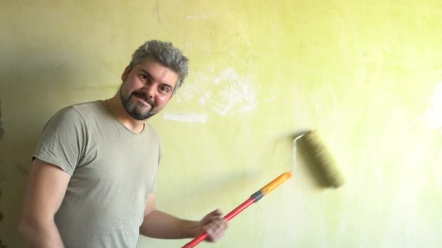A bearded man paints a wall. Good mood. A smile on his face. Repair work in the room. Actor. Frick. Clown.