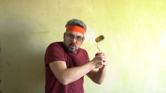 Bearded man against the wall. Good mood. Engaged in martial arts. In the hands of a construction roller for wallpaper. Repair work in the room. Frick.