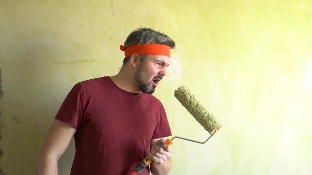 Bearded man against the wall. Sings in a construction roller. Performance of the artist. Musical number. On the head is an orange ribbon. Repair work in the room. Frick. Retro style. Green wall. Rock-
