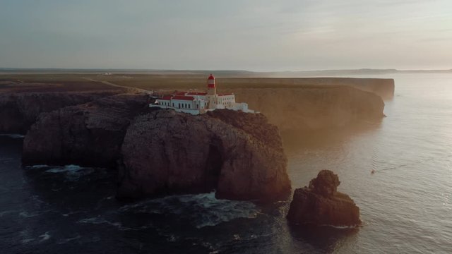 Magnificent aerial view of the lighthouse Cabo de S&atilde;o Vicente on a cliff surrounded by clean blue Atlantic ocean in Sagres, Portugal on sunrise. A boat is passing by.
