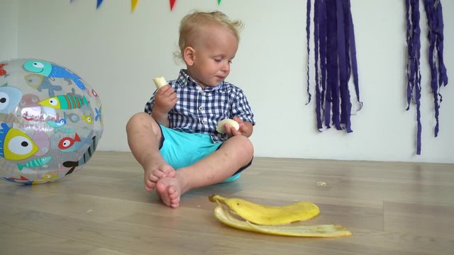 Cute baby child eating banana sitting on floor. Handsome kid with banana in hand