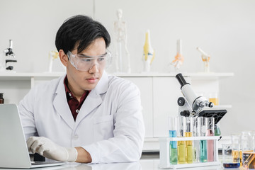 A male scientist with black hair wearing white coat and protective glassware sitting with laptop looking at a microscope in white laboratory room with test tube and solutions.