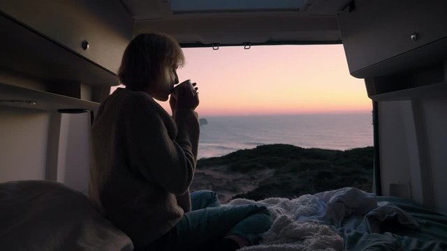 Young woman sitting and relaxing in her Camper van drinking hot tea / coffee. Van is parked on a cliff with a beautiful view on the sunset/sunrise. Epic life and travelling the world.