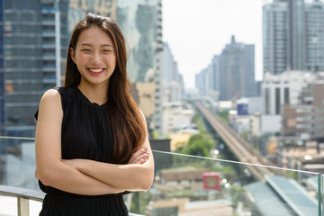Happy young beautiful Asian businesswoman smiling with arms crossed against view of the city