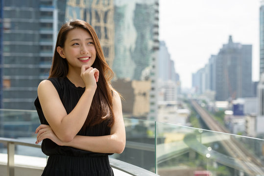 Happy young beautiful Asian businesswoman thinking against view of the city