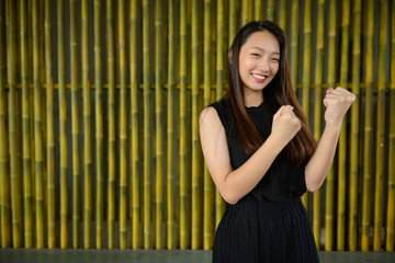 Happy young beautiful Asian businesswoman against bamboo fence