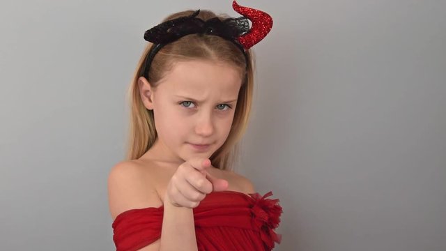 Portrait of a cute little blonde girl in a red T-shirt with devil horns pointing to the screen with her finger meaning you on a gray background. Concept of advertising and motivation.