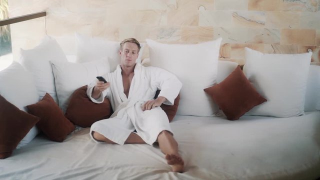 blond guy in bathrobe switches on TV on comfortable bed