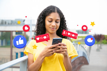 Happy excited Latin girl using smartphone. Young black woman in yellow casual shirt holding mobile phone, texting message, chatting online with like and comment notification icons. Wi-Fi concept