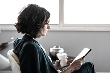 Businesswoman reading news in office lounge. Business woman sitting in armchair, drinking coffee, holding tablet, and looking at screen. Wireless connection concept
