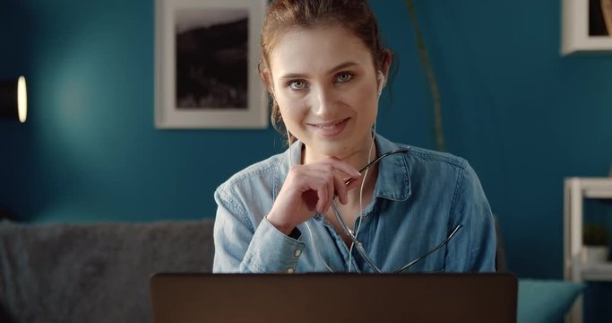 Portrait of pretty young woman with hair knot smiling and looking at camera while sitting at desk with laptop. Charming girl in casual clothing enjoying free time at home.