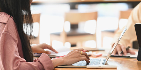 Cropped image of creative woman in pink long sleeve working on computer laptop and writing on notebook while sitting at the modern wooden table with comfortable cafe as background.