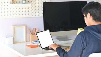 Cropped shot of smart man holding a coffee cup in hand while typing white blank screen computer laptop and sitting at the modern working table with comfortable workplace as background.