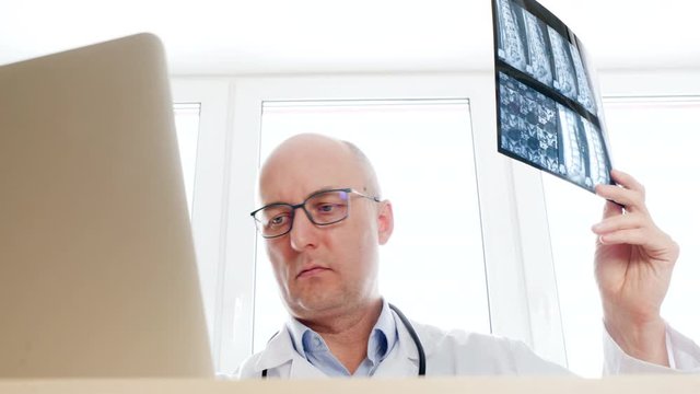 Surgeon looking backbones x-ray front laptop screen in medical office. Male doctor examining x-ray and working on computer. Medical worker looking xray picture