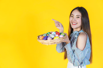Asian young woman pointing finger to blank copy space and looking at camera, hold basket filled easter eggs, toothy smiling happy face. Holidays and people concept isolated on yellow color background