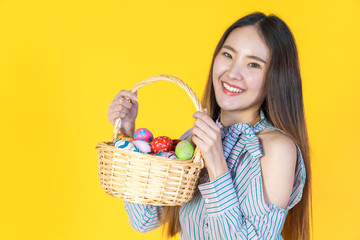 Attractive young Asian woman standing, looking at camera, hold basket filled easter eggs, toothy smiling happy face. Holidays and people concept isolated on blank copy space, yellow color background.