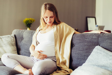 Peaceful expectant mother caressing baby bump. Young pregnant woman sitting on couch, touching belly. Pregnant woman at home