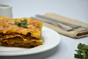 A piece of lasagna on a white plate. White background with kitchen utensils.
