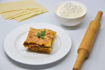 A piece of lasagna on a white plate. White background with pasta sheets, flour, and a pin.