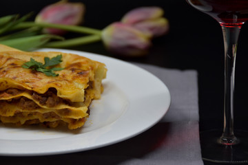 A slice of lasagna on a white plate. Black background with tulips and a glass of red wine.