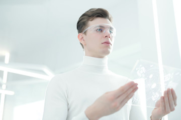 Serious handsome young man in smart goggles working with ultramodern transparent device in office