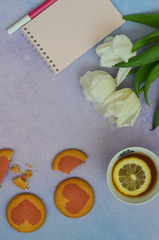 Top view of a cup of tea with lemon, cookies, a notebook with a felt-tip pen and white tulip flowers on a purple background. Space for text
