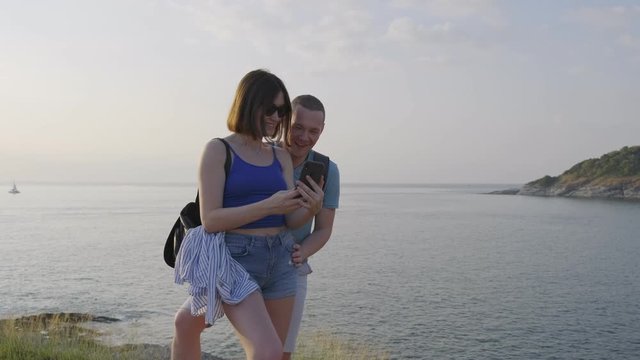 Young couple near the sea, lake browsing photos and selfie on the phone. Romantic man and woman laugh and wonder. 4k footage