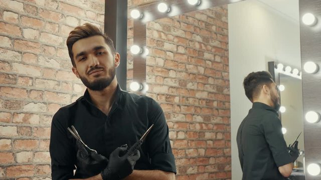 Handsome barber holding scissors in crossing hands front camera in beauty salon. Portrait smiling hairdresser with scissors posing front camera in barbershop