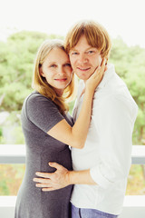 Happy expectant parents hugging and posing outside. Man and pregnant woman standing on balcony, embracing each other and looking at camera. Expecting baby concept