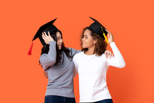 Portrait of two happy mixed-race women graduating holding there graduation caps looking at each other in orange isolated studio background