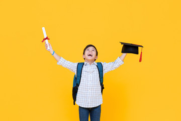 Happy graduate boy with academic cap smiling and raising hands celebrating graduation day isolated on yellow background