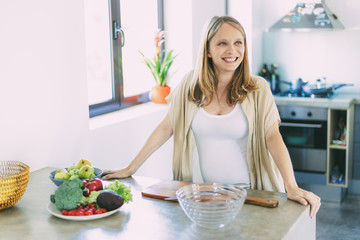 Obraz na płótnie Canvas Joyful expectant mother cooking salad in kitchen. Young pregnant woman standing at table with bowls of vegetables, smiling, looking away. Pregnancy and nutrition concept