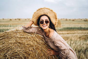 Smiling woman in sunglasses with bare shoulders on a background of wheat field and bales of hay - obrazy, fototapety, plakaty