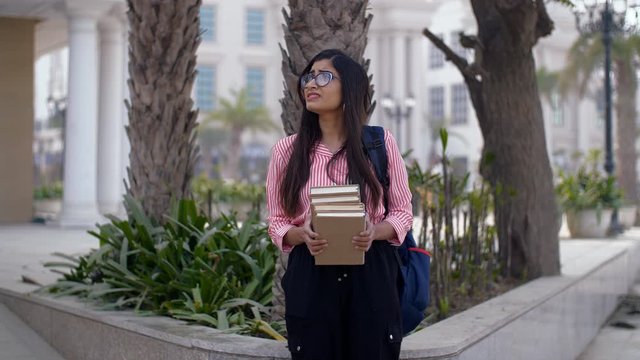 Young college girl holds a stack of books and frowns at them - unhappy emotions. Frustrated Indian female wearing a backpack  holding a pile of books while standing outside the college campus - rej...