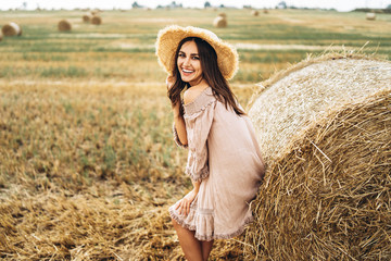 Naklejka na ściany i meble Beautiful young girl with long hair in sunnglasses and straw hat posing on a wheat field near hay bales. Happy brunette in summer dress